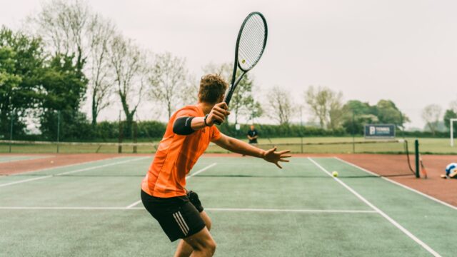 man in orange shirt and black shorts holding black and white tennis racket